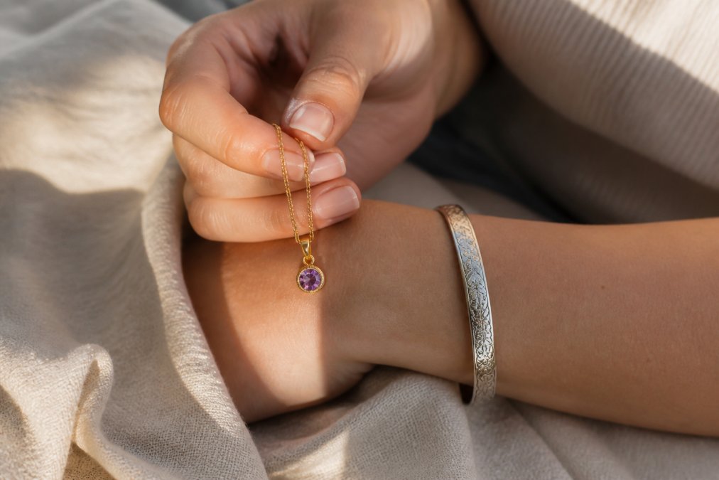 Close-up of a woman's wrist and hand gently holding a gold birthstone pendant necklace and an engraved silver cuff bracelet against a soft linen cloth, with warm natural window light catching the metal finishes