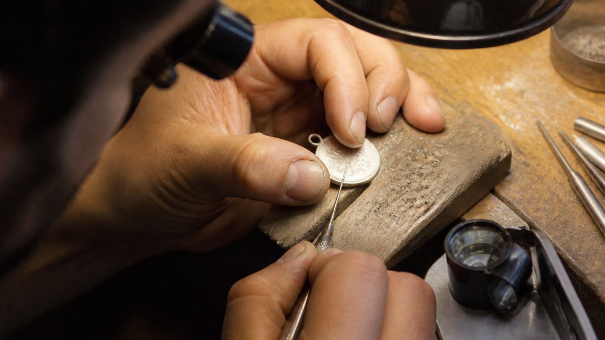 Close-up of a jeweler's hands guiding a fine engraving tool across a silver pendant on a workbench, warm lamp light, scattered metal shavings and a loupe nearby