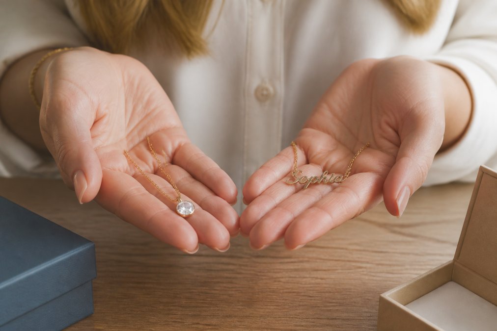 A woman's hands holding two necklaces side by side: one simple generic store pendant and one custom name necklace with a cursive design, soft natural light on a wooden table