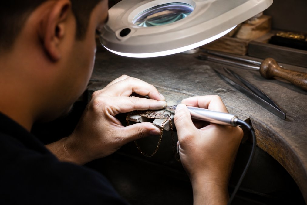 Close-up of a jeweler's hands carefully engraving a personalized name onto a custom necklace using precision hand tools in a well-lit workshop with magnifying lamp