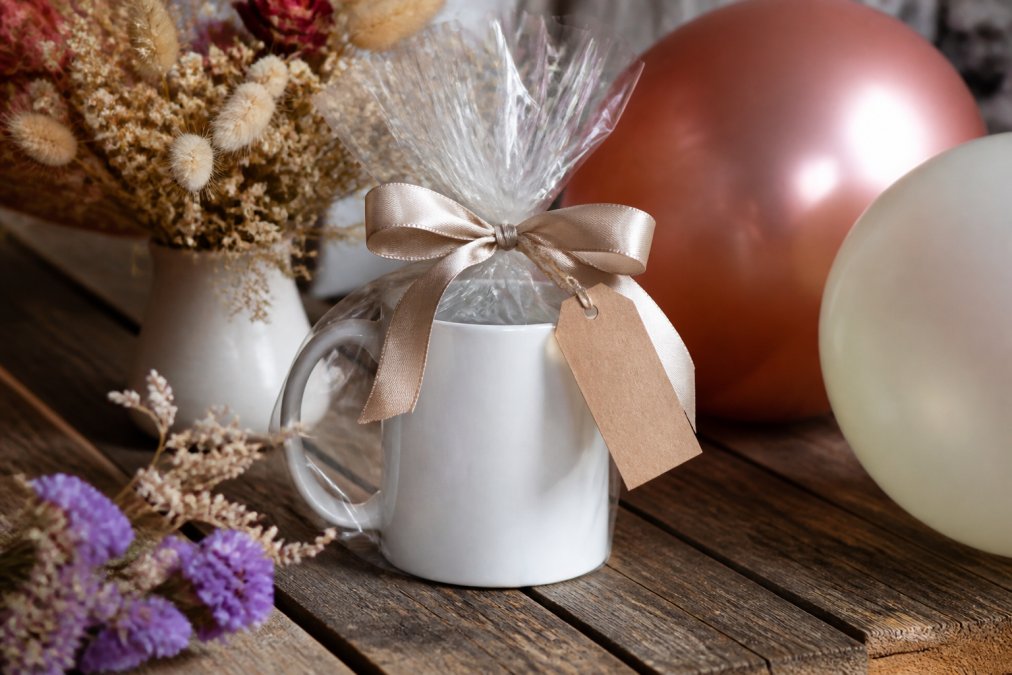 A beautifully wrapped white ceramic mug tied with a satin ribbon and a handwritten gift tag, placed on a rustic wooden table surrounded by birthday balloons and dried flower decorations in warm ambient lighting