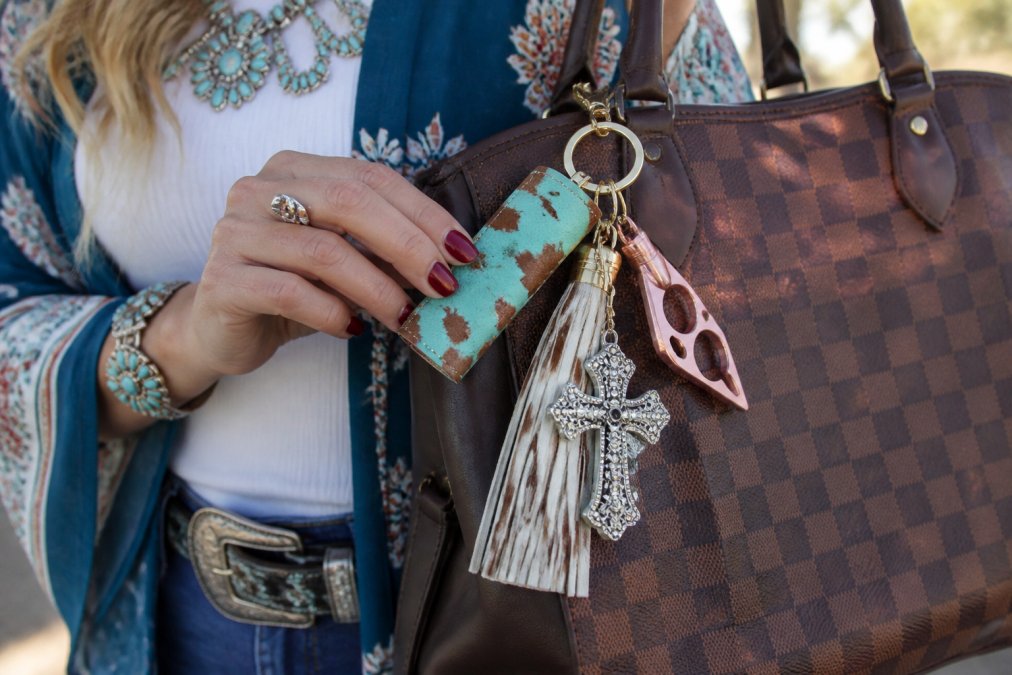 Close-up of a woman's hand holding a stylish self-defense keychain with Western-inspired design attached to a designer handbag, showing how the accessory complements her outfit in natural daylight