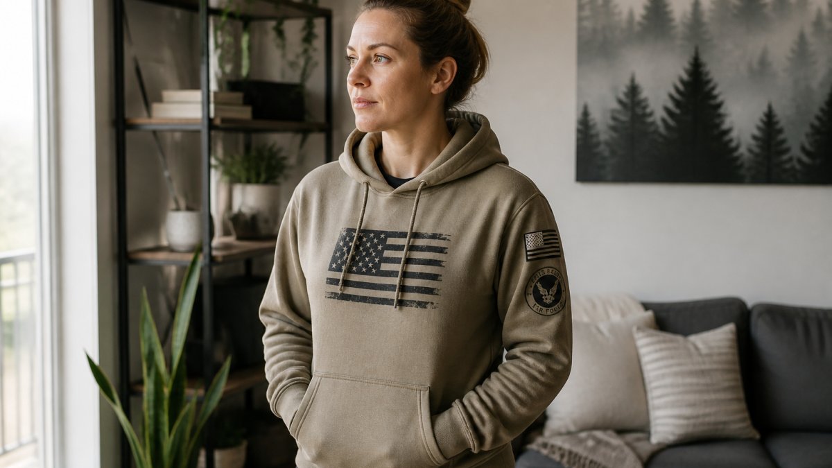 Female veteran wearing well-fitted patriotic hoodie with branch insignia in neutral color palette, standing confidently in modern home setting with natural lighting
