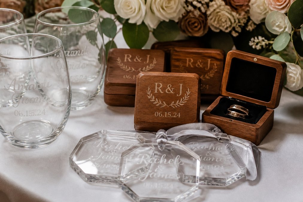 Close-up of various engraved wedding favors displayed on white linen, including personalized wine glasses with names and dates, custom wooden boxes with initials, and acrylic keepsakes with couple's names and wedding date visible in elegant script