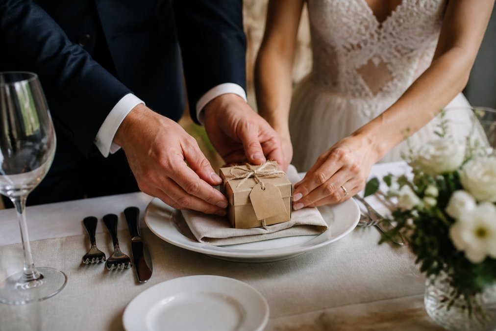 Close-up of a bride and groom's hands placing a small personalized gift box with a handwritten tag onto a reception table setting with soft window light