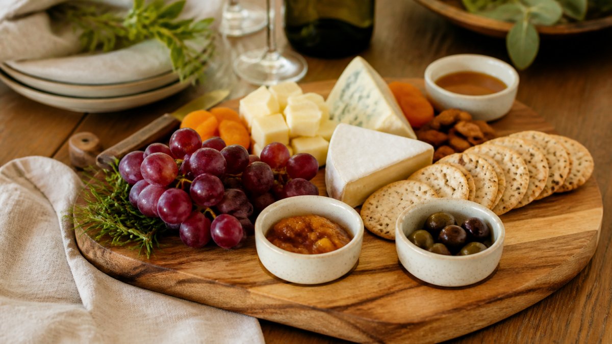 A close-up of an engraved acacia wood charcuterie board with ceramic dipping bowls, styled with cheese, crackers, and grapes on a rustic table with warm lighting