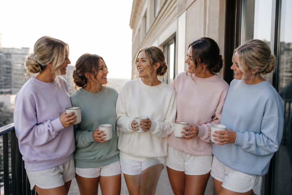 A bridal party of five women wearing coordinated personalized sweatshirts in soft pastels, standing together laughing on a hotel balcony during morning preparations