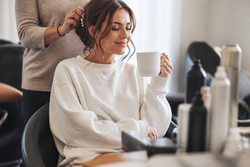 Close-up of a bride in a cozy oversized embroidered sweatshirt with her initials, sipping coffee while getting her hair done, with wedding day styling products visible nearby