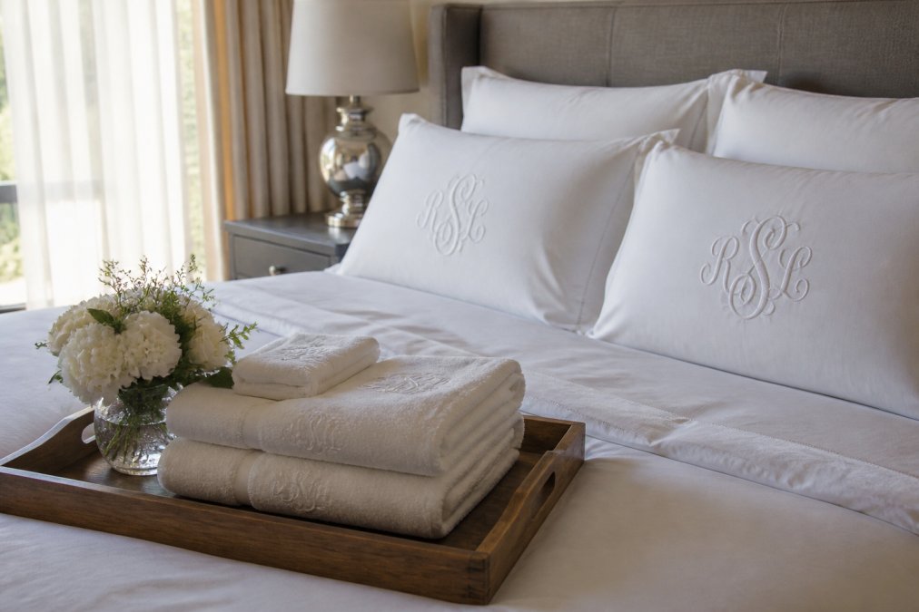 A neatly made bed with crisp white monogrammed linen pillowcases and a folded monogrammed bath towel set arranged on a wooden tray beside a small vase of fresh white flowers, soft morning light through sheer curtains