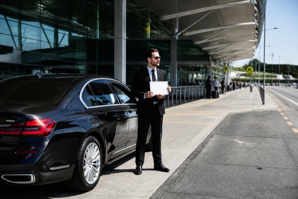 A professional chauffeur in a dark suit standing beside a polished black luxury sedan at an airport kerbside drop-off zone, holding a printed name sign, with the glass terminal entrance and departure signage visible in the background under bright midday light