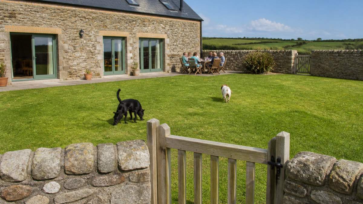 A fully enclosed lawned garden at a stone barn conversion with a low stone wall and secure gate, two dogs exploring the grass while a family sits at an outdoor table in the background