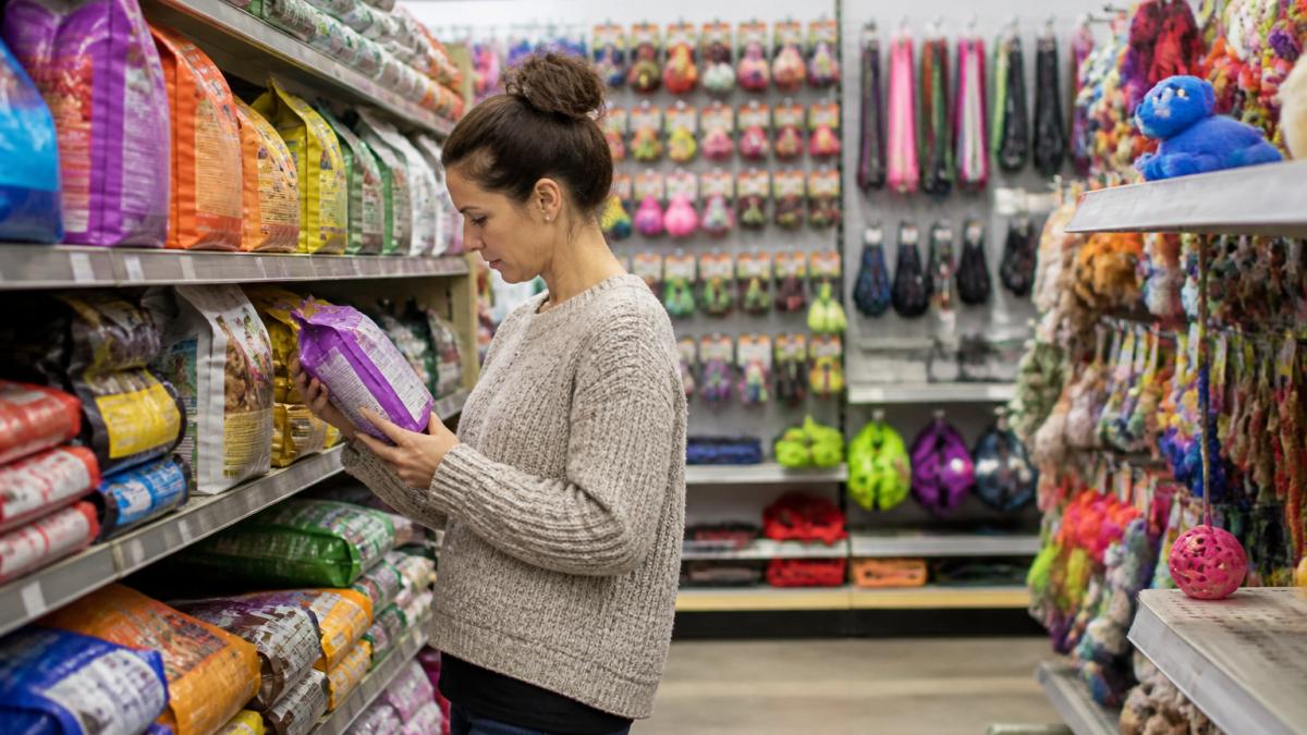 Pet owner browsing shelves of colorful pet food bags, toys, and accessories in a bright retail environment, examining product labels and packaging carefully