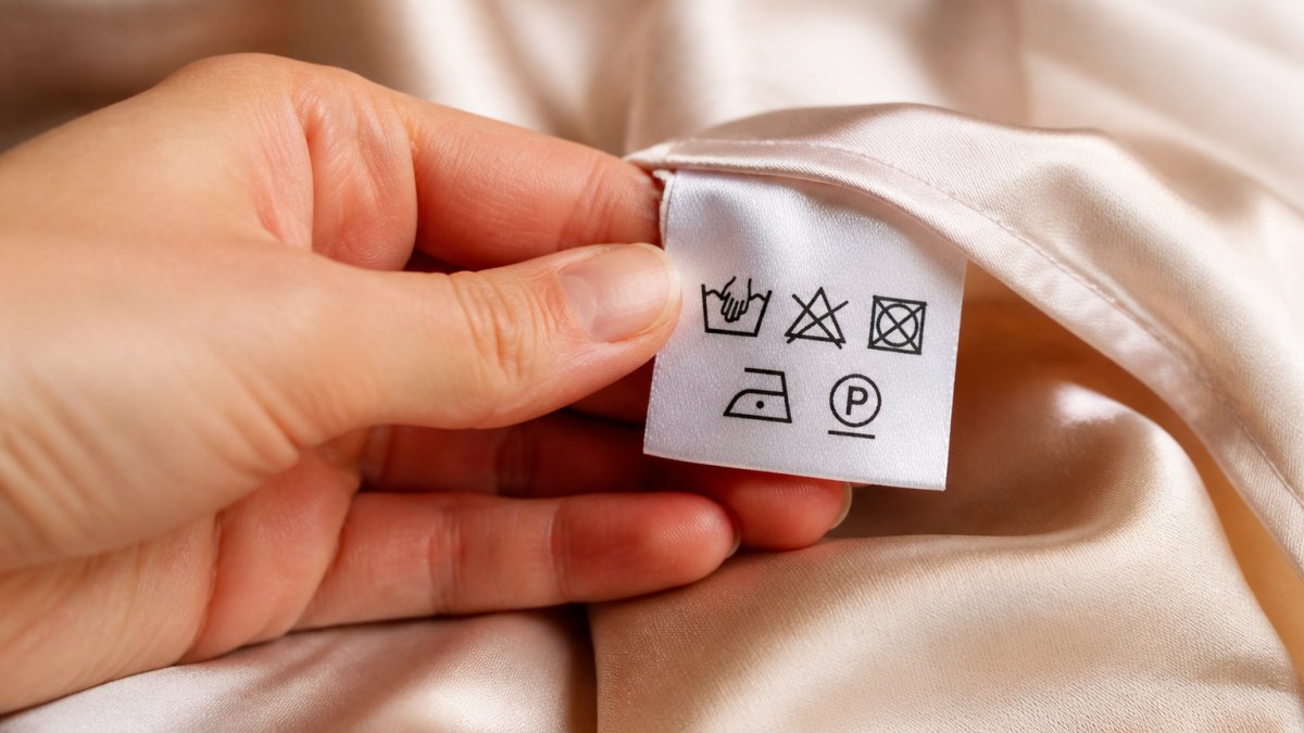Nahaufnahme einer Hand, die ein weißes Pflegeetikett an einem eleganten Seidenkleid untersucht, mit deutlich sichtbaren Pflegesymbolen in Schwarz
