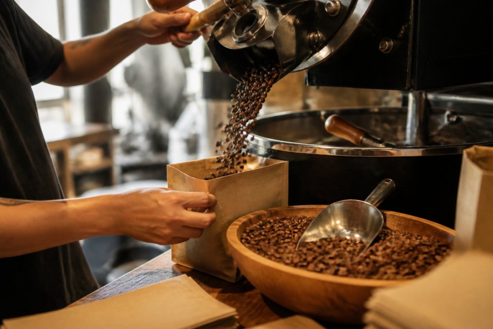 Close-up of freshly roasted coffee beans cascading into a kraft paper bag at a specialty roastery workbench, with a handwritten roast date label and wooden roasting equipment visible in soft afternoon light