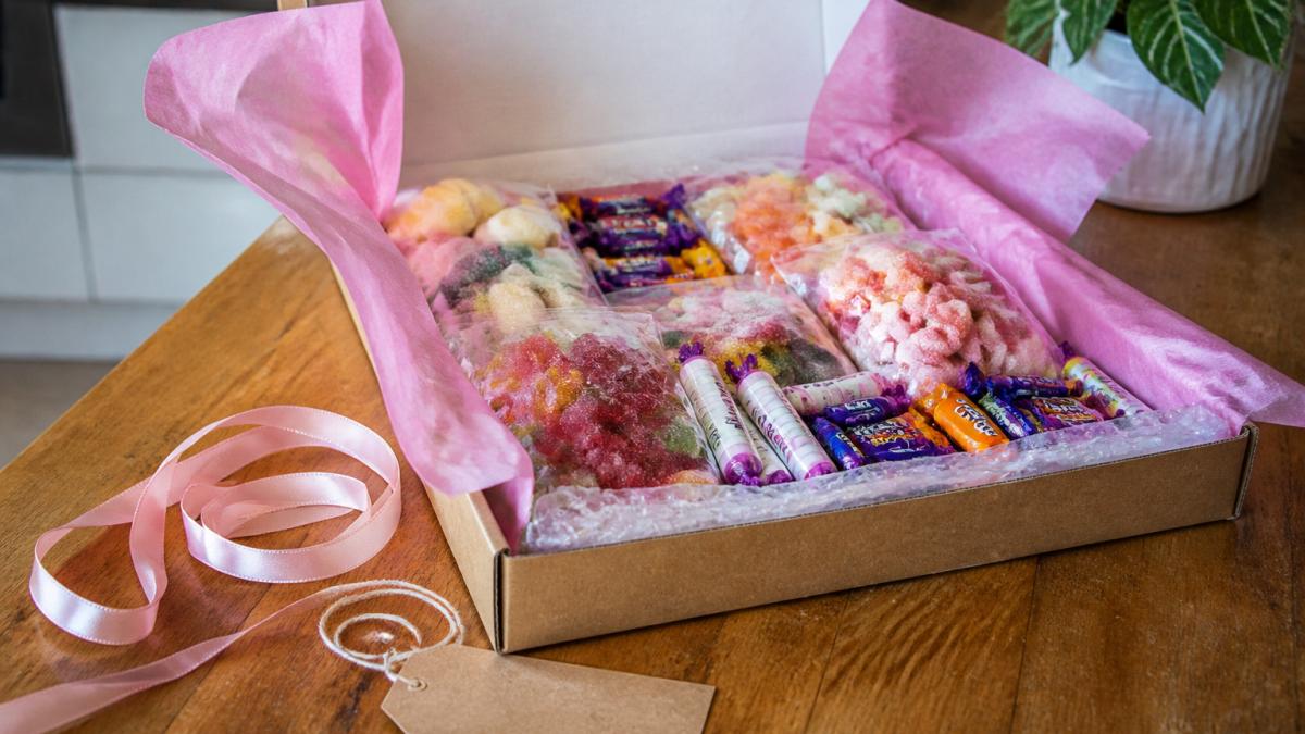 A colourful pick and mix sweet gift box open on a kitchen table, filled with wrapped retro sweets and jelly favourites, with ribbon and a handwritten gift tag beside it