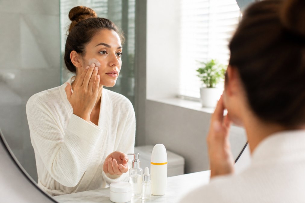 Person applying sunscreen to face with fingertips in morning bathroom routine, with skincare products and sunscreen bottle visible on marble counter beside mirror