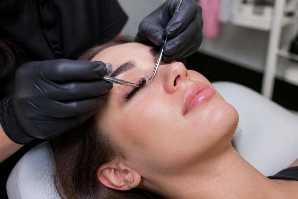 Close-up of a lash technician in gloves applying individual eyelash extensions with fine tweezers to a client lying on a treatment bed under bright studio lighting