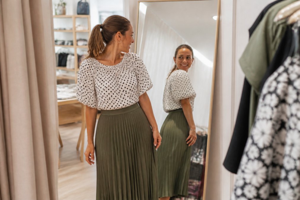 A woman in her thirties trying on a flowy olive pleated midi skirt and a polka dot puff sleeve blouse in a bright boutique fitting room, smiling at her reflection in a full-length mirror