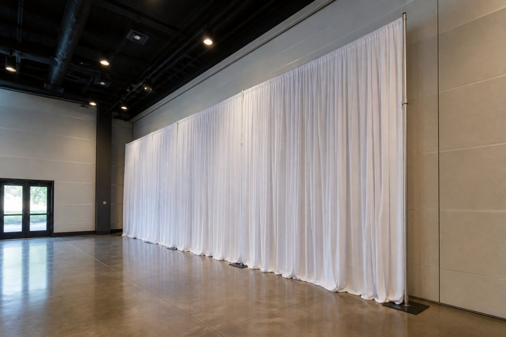 Professional event space with tall pipe and drape backdrop installed, showing telescopic uprights extended to ceiling, weighted base plates anchoring the system on polished floor, and draped fabric creating a professional seamless backdrop
