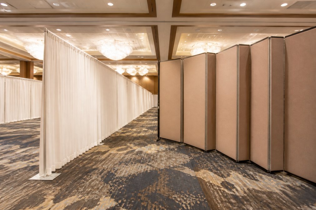 Wide shot of a hotel ballroom divided by elegant white pipe and drape on the left, with a rigid accordion-style fabric room divider visible on the right side of the frame, warm ambient lighting