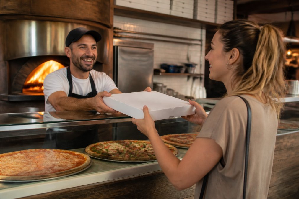 A server hands a fresh box of pizza Warwick style to a customer inside a cozy, wood-fired family pizzeria.