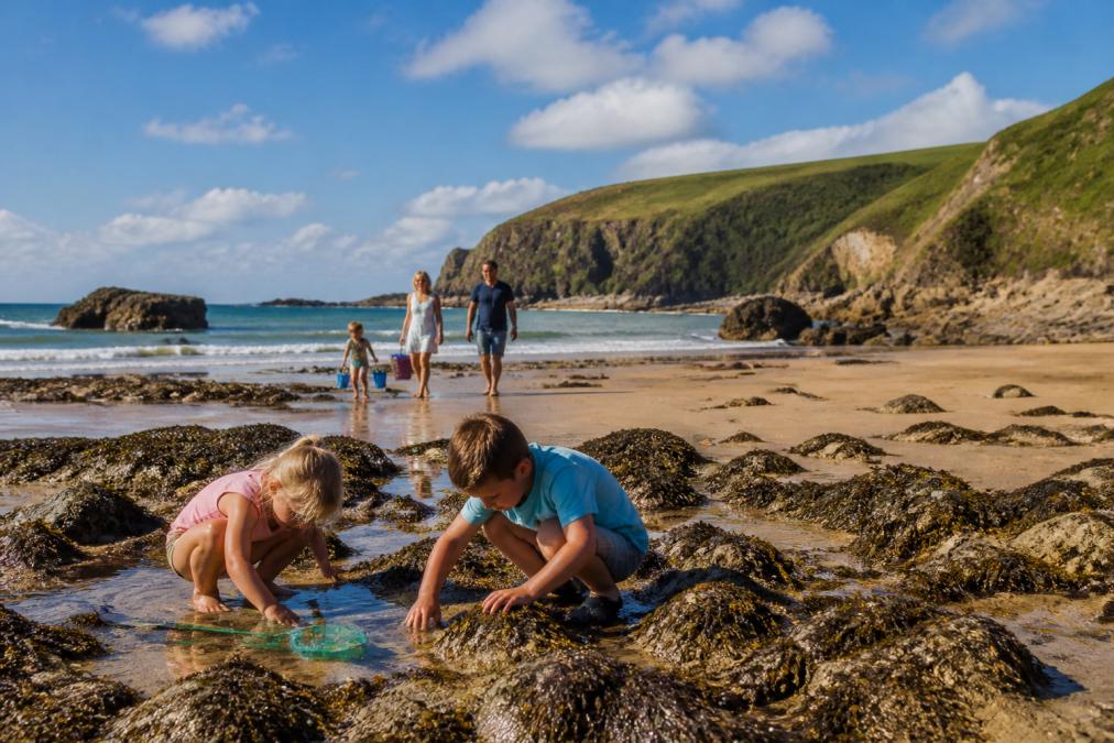 Family with young children playing on sandy beach with rock pools visible, dramatic cliffs in background, and coastal path views on a sunny afternoon