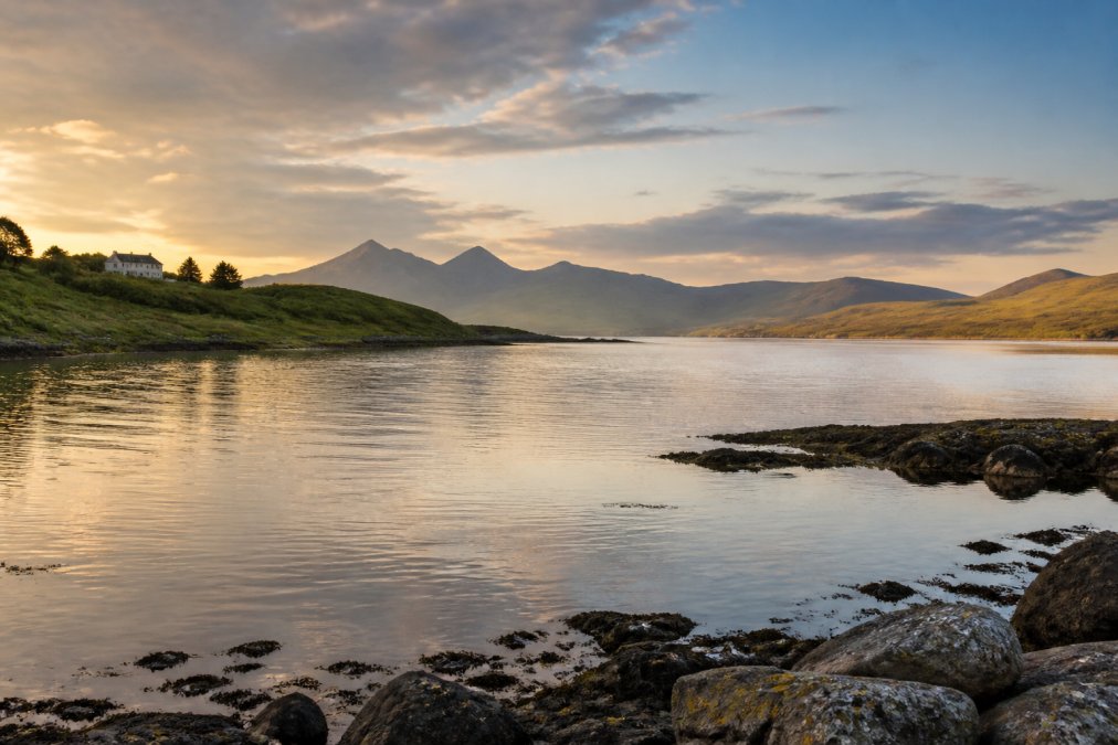 A serene view of the Isle of Skye coastline at golden hour, calm loch water in the foreground with rolling green hills and distant Cuillin mountains under a soft evening sky