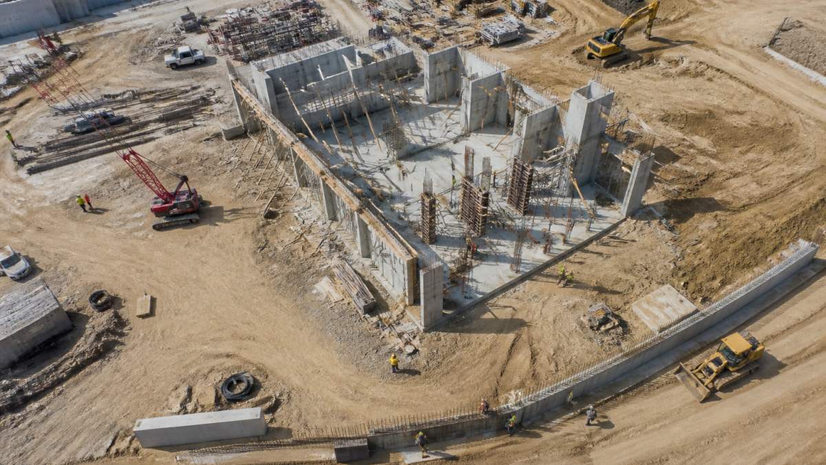 Aerial view of an active construction site with multiple pieces of equipment, workers in safety gear, and partially completed concrete structures visible from above, captured during midday with clear shadows showing depth and progress