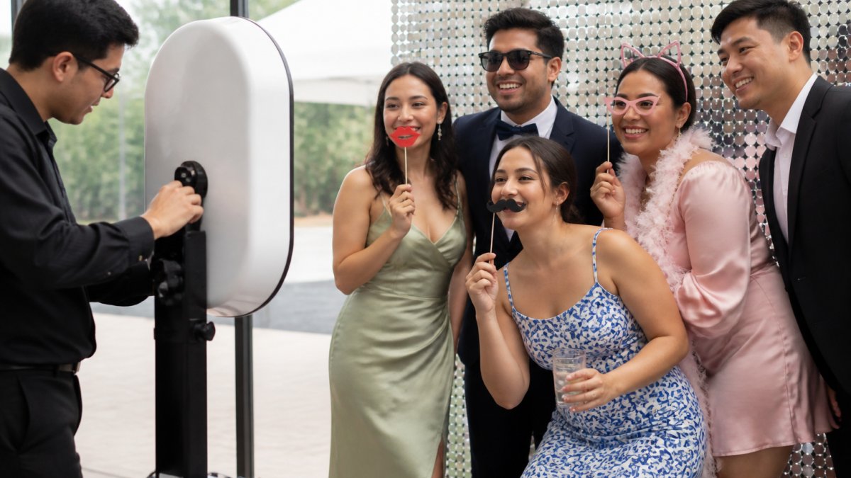 Wedding guests smiling and laughing while posing with props in front of a photo booth backdrop, with a trained attendant adjusting the camera setup
