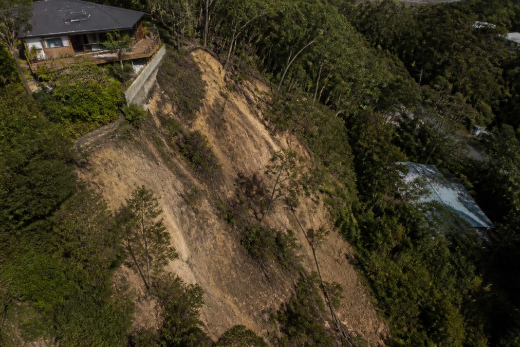 Aerial view of a steeply sloped residential block showing visible contours, exposed soil on the slope, and the natural gradient of the land with trees and vegetation