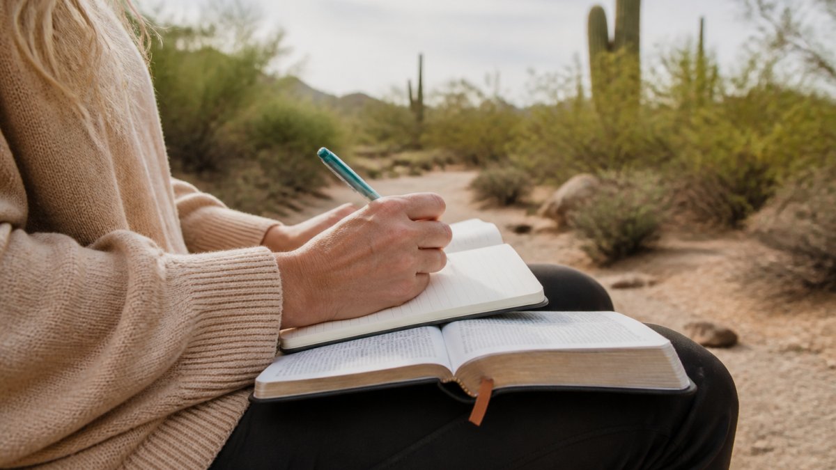 Person sitting on a desert trail with a journal and Bible during a quiet spiritual retreat at sunrise.