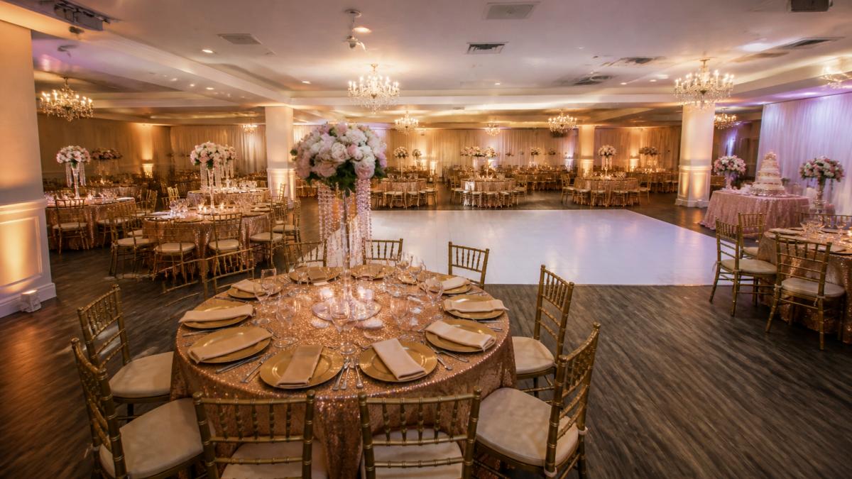 An elegantly decorated banquet hall set for a quinceañera with round tables, draped linens, warm up-lighting, and an open dance floor, showing the scale of a 150-guest reception space