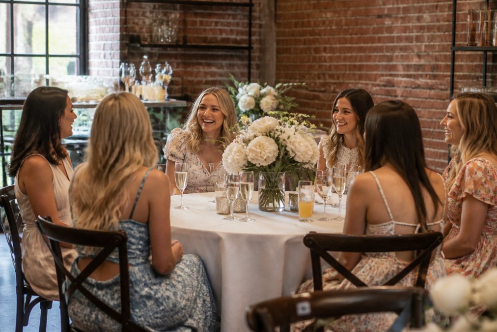 A warmly lit coffee shop event space set up for a small intimate bridal shower, with white floral arrangements on round tables, champagne glasses catching the light, and six women in casual dresses chatting and laughing in a cozy, brick-walled room