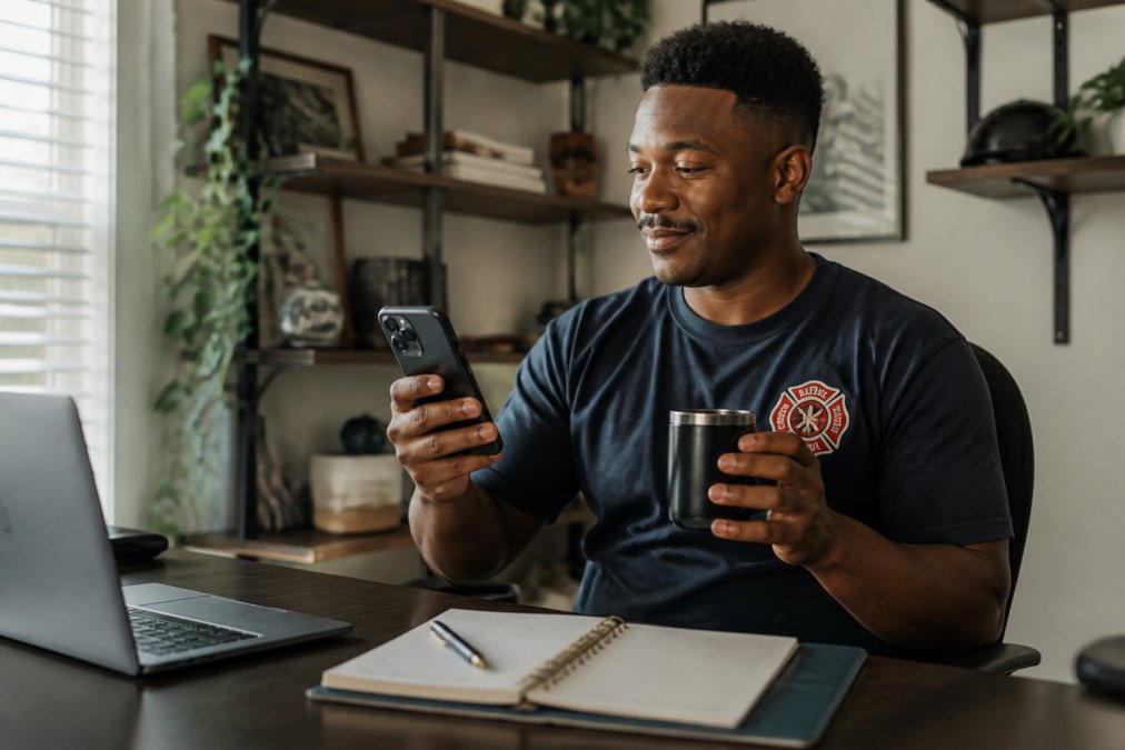 First responder or shift worker reviewing a calendar on their phone while holding coffee, planning travel dates with a relaxed expression in home office setting