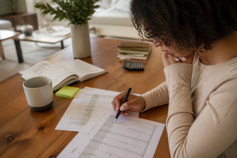 Person sitting at a desk with a printed budget spreadsheet, a pen, and a laptop open, reviewing event cost categories with focused concentration
