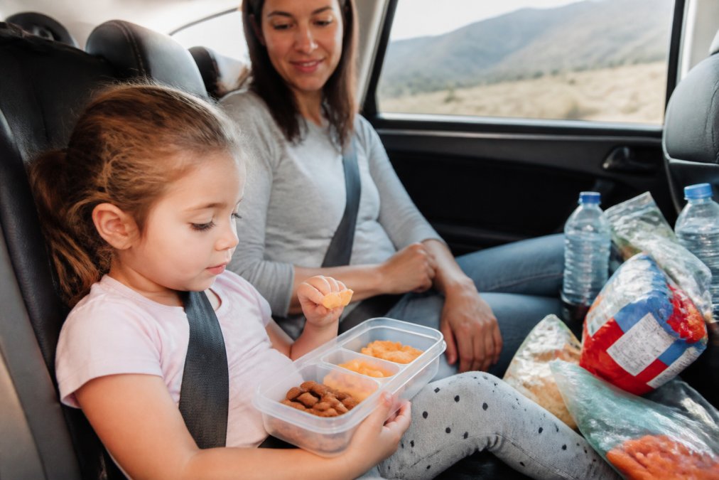 Parent and child sitting in a car during a road trip, child eating from a labeled plastic snack container, with various packaged snacks and water bottles visible on the car seat, natural daylight streaming through windows