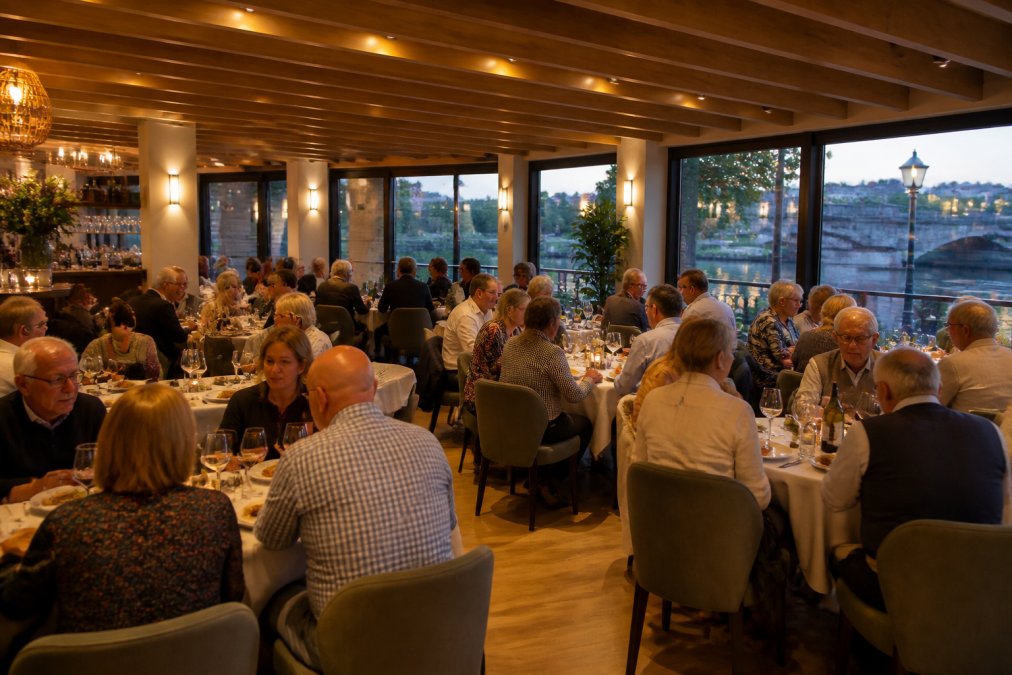 A warm, intimate dining space with 40 guests seated at round and rectangular tables, enjoying food and conversation, with soft ambient lighting and riverside views visible through large windows