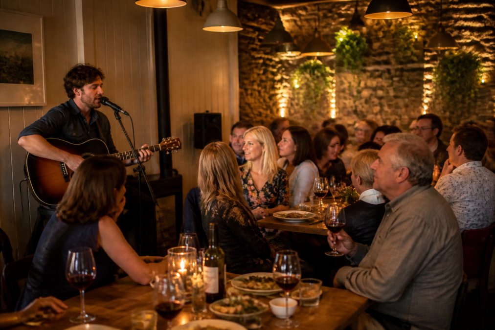 A musician with an acoustic guitar performing in a cosy venue setting with guests gathered around tables, enjoying food and conversation, with warm ambient lighting and intimate atmosphere