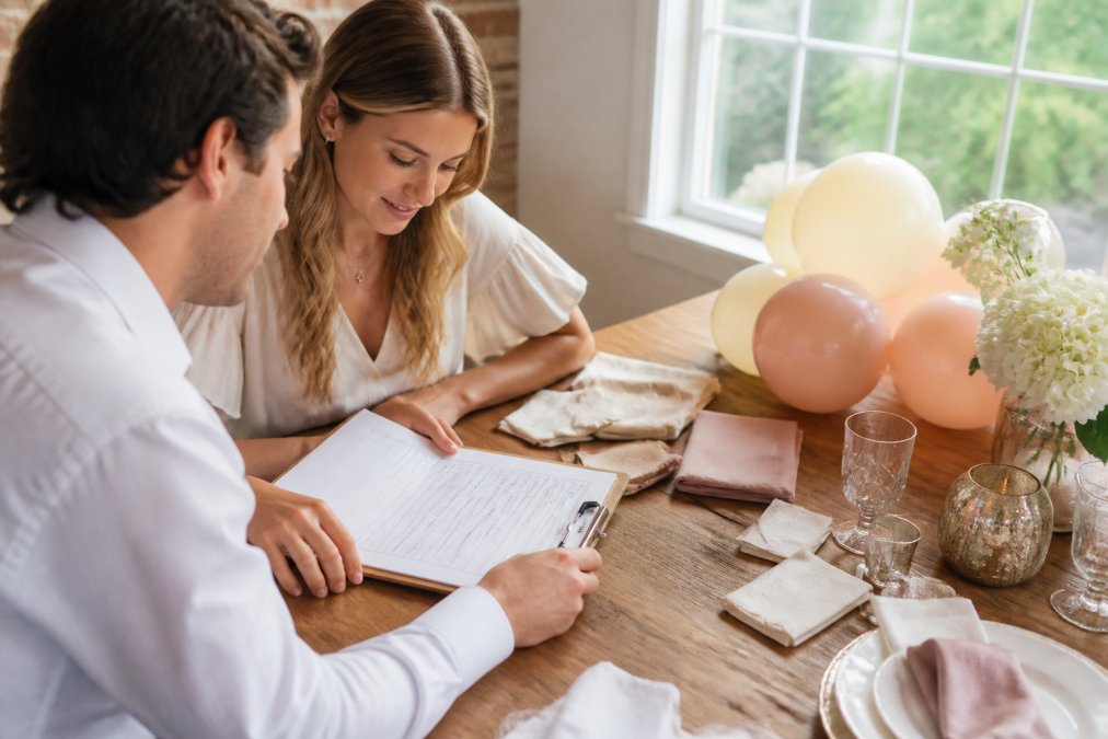 A couple reviewing a wedding planning checklist on a clipboard at a wooden table covered with fabric swatches, balloon samples in ivory and blush, and event décor items, with natural window light illuminating the scene