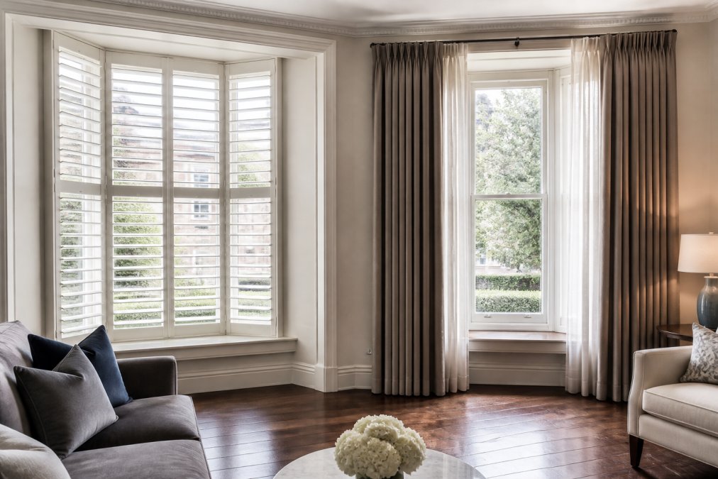 A bright living room in a period London home with tall bay windows, one side fitted with white plantation shutters with tilted louvres, the other side dressed with full-length lined curtains, natural daylight filtering through both