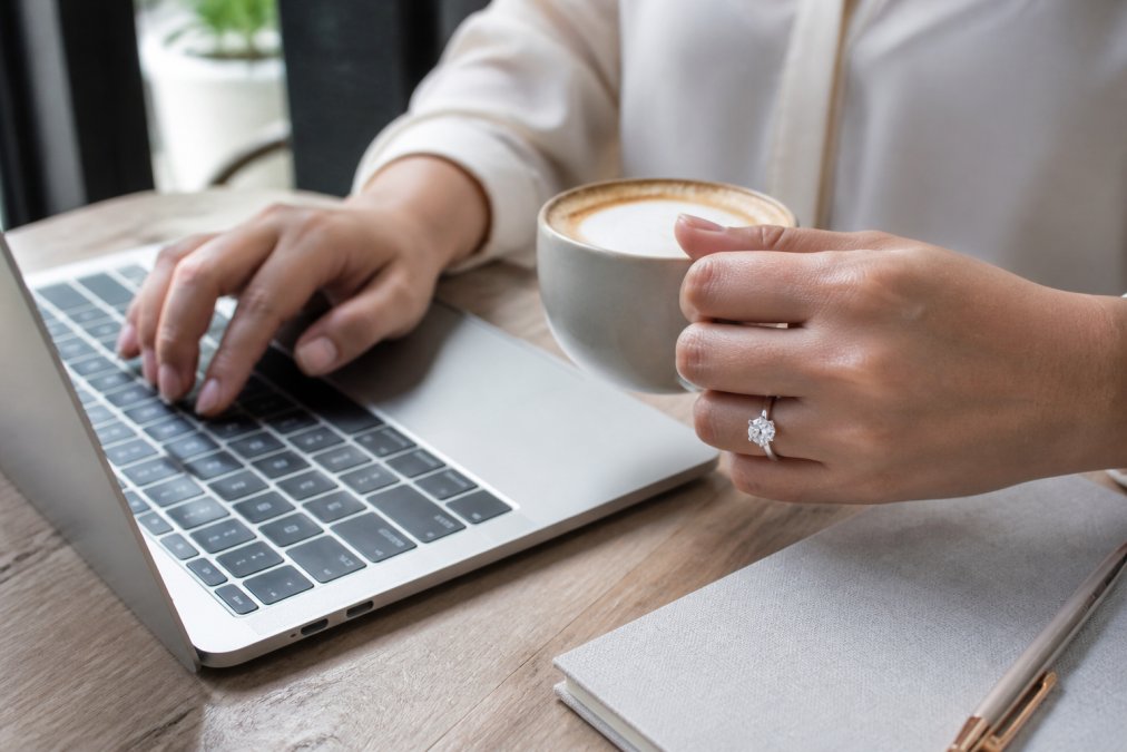Woman's hand wearing a platinum engagement ring during everyday activities, typing at a desk, holding a coffee cup, in natural daylight, showing the ring's lustre and how it catches light