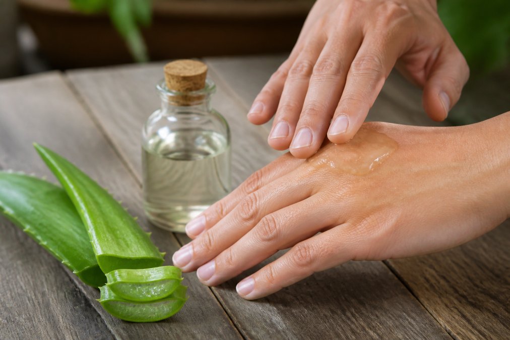 Close-up af hænder, hvor transparent Aloe Vera-gel påføres med fingrene, med friske aloe-blade og en lille glasflaske på en naturlig træoverflade