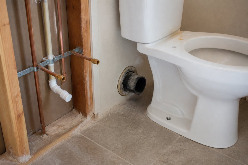 Close-up of a bathroom toilet installation showing visible rough plumbing connections and drainage lines with new fixture ready for connection