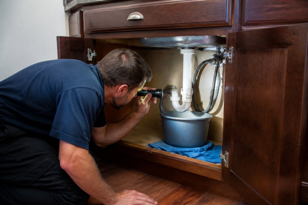 Homeowner using a flashlight to inspect water stains under a sink for plumbing maintenance.
