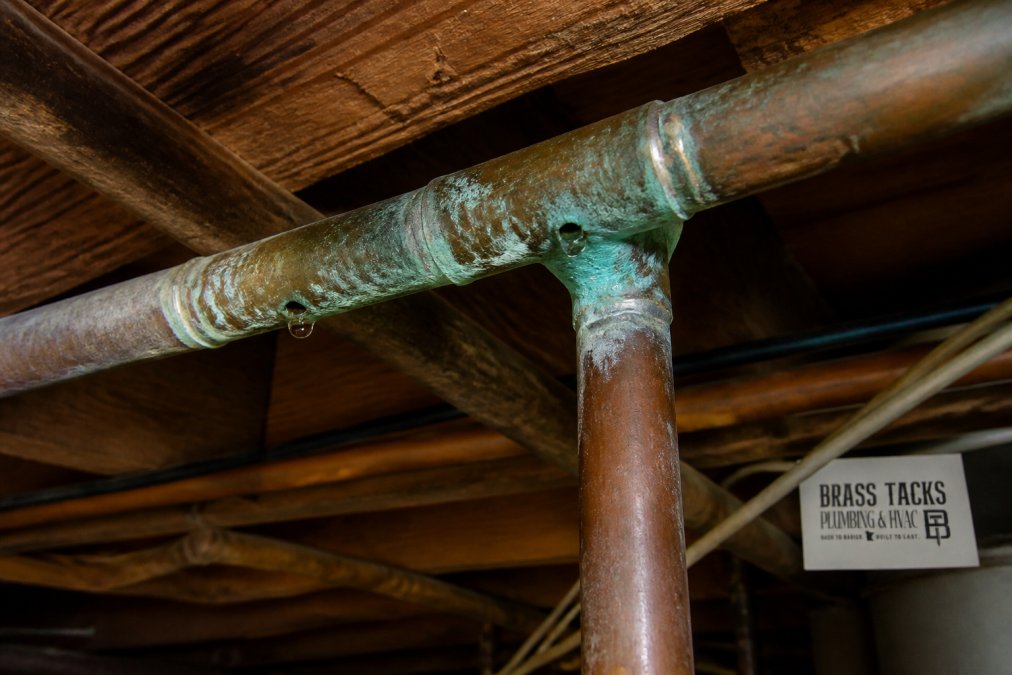 Close-up of corroded copper pipes with visible pinhole leaks and green patina staining on basement ceiling piping near a junction