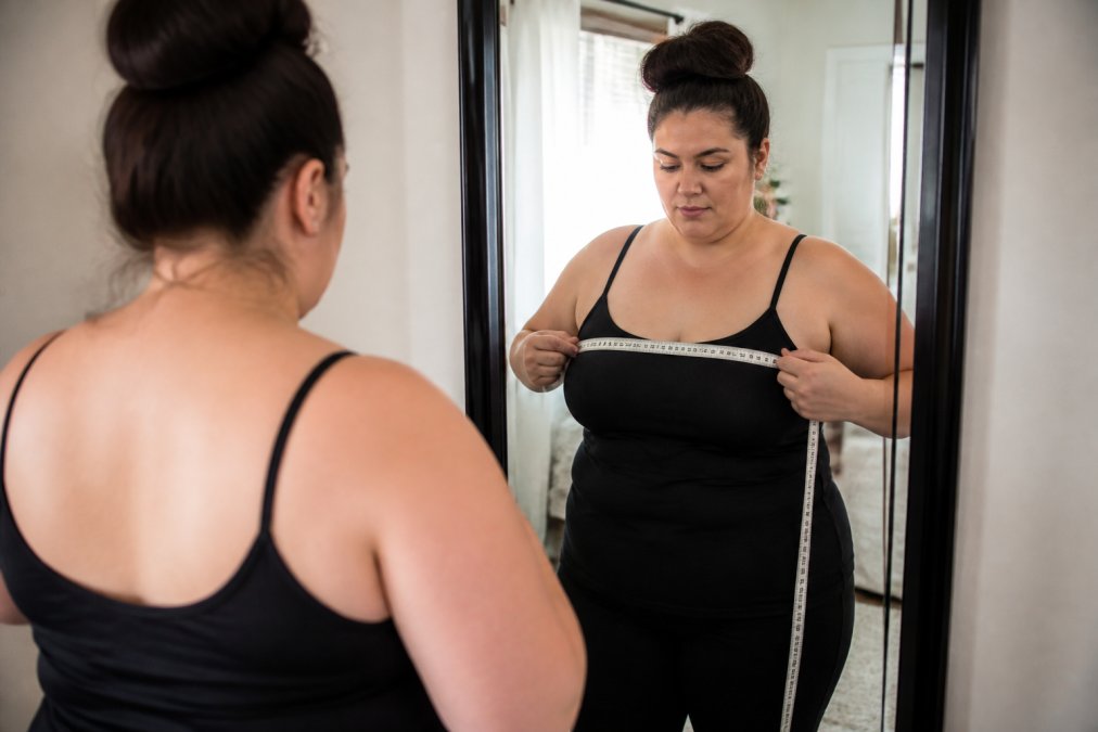 Woman using a soft measurement tape around her bust while standing in front of a mirror, demonstrating proper snug placement with clear visibility of the tape measurement