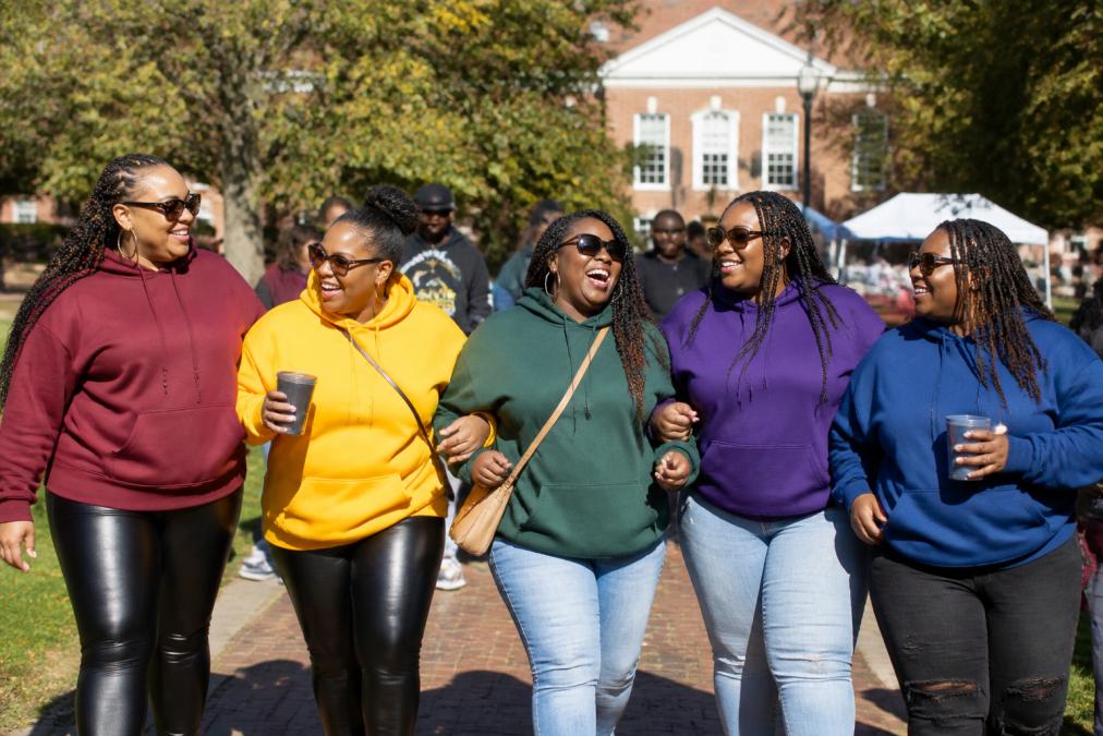A group of plus size HBCU alumni and students wearing oversized collegiate hoodies in school colors, laughing and walking together on a sunny campus quad during homecoming weekend