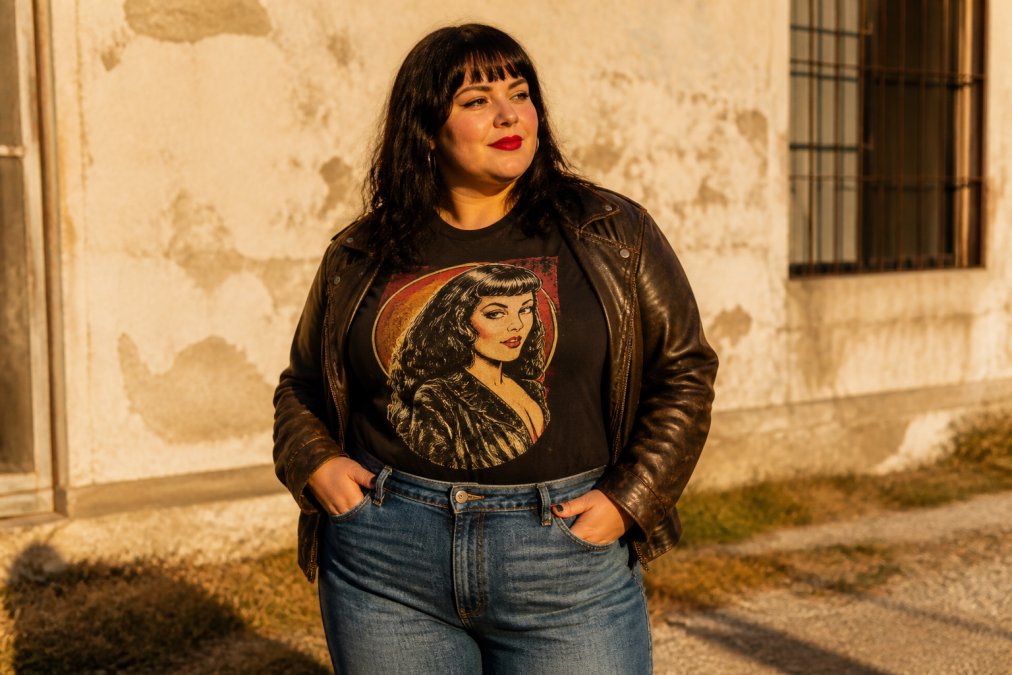 Person wearing a retro graphic tee styled with vintage-wash denim jeans and a vintage leather jacket, standing outdoors with natural sunlight highlighting the graphic design and fit across the torso and shoulders