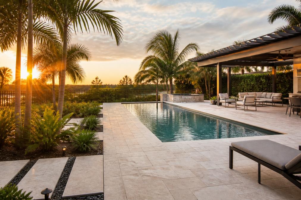 A modern South Florida backyard at golden hour featuring a custom-built rectangular pool with clean travertine patio pavers, tropical landscaping, and an outdoor lounge area with a pergola