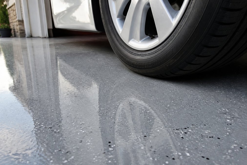 A close-up of a residential garage floor coated in glossy gray polyaspartic, with a car tire resting on the surface, showing the coating's resistance to hot tire pickup and UV exposure